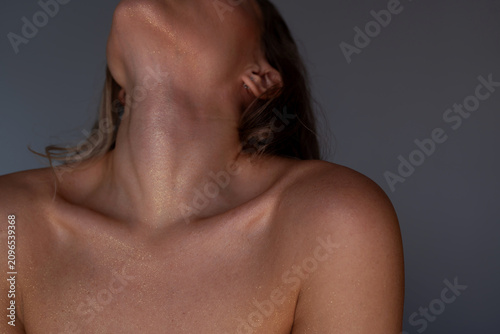 Close-up of young woman neck and collarbones with golden shimmer on glowing skin, sensual beauty concept with soft studio light on a grey background