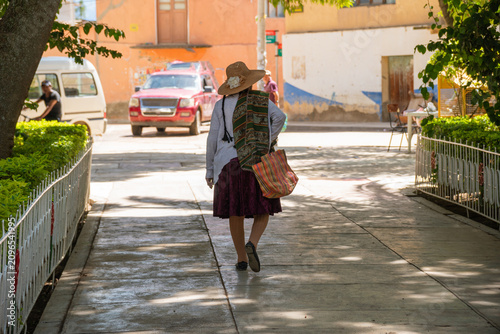 Rear view of indigenous bolivian woman walking on town square street wearing traditional clothes and straw hat