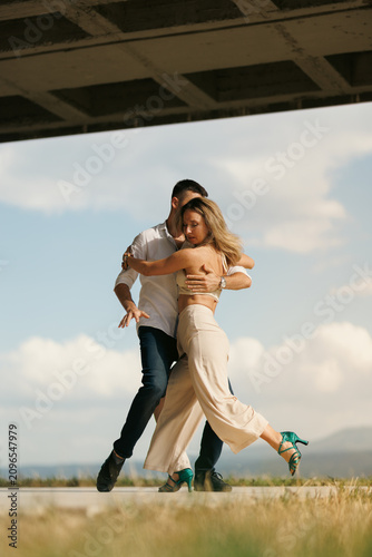 Couple dancing tango outdoors under a bridge, embracing passionately in a sunlit romantic dance