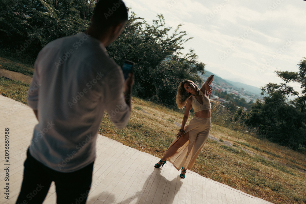 Obraz premium Photographer capturing a female model dancing and posing during an outdoor fashion photo shoot on a sunlit hillside with city view