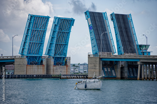 Bild auf Leinwand Blue bascule drawbridge raised above a calm harbor as a motorboat and sailboat pass beneath in St