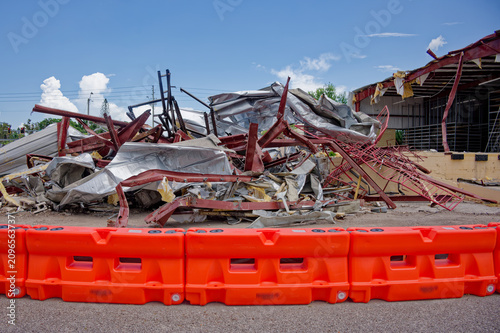 Collapsed metal buildings and twisted red steel beams sit behind bright orange safety barriers at an urban demolition site, with rubble and debris under a clear blue sky suggesting danger and cleanup.