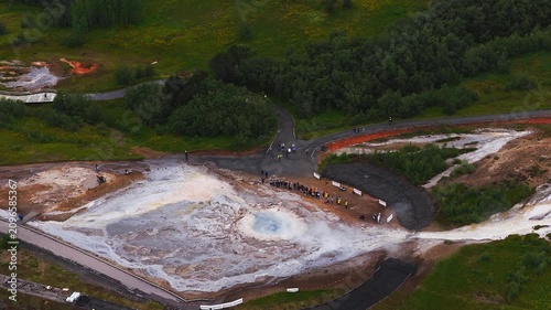 Aerial view shows a vivid Strokkur geyser pool, white and orange terraces, sinter channels, and visitors on paths in Haukadalur, Iceland, as steam rises and water flows.