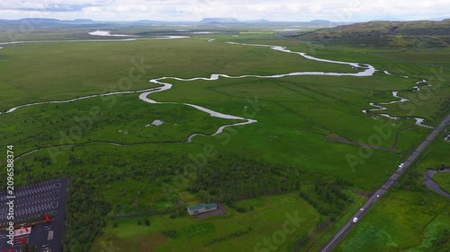 Aerial view of Icelandic lowland with sinuous river, braided streams, flat topped mountains, highway with camper vans, parking area, and a lone building in soft light.