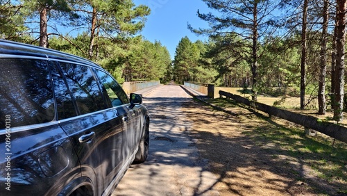 A short stop during a spring trip. The car is parked on an old asphalted road with pits in front of a bridge with metal fences. A river flows nearby, and beyond it is a forest. Sunny spring weather
