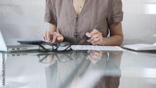 Obraz na płótnie Businesswoman accountant, wearing brawn dotted blouse, is using a calculator to crunch numbers and jotting down meticulous notes, at sleek glass desk in a contemporary office