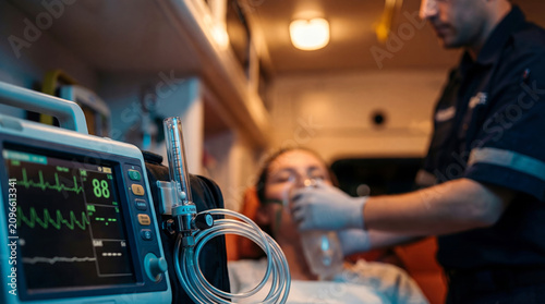 Close-up of a medical monitor and oxygen flow meter inside an ambulance while a paramedic assists a patient wearing an oxygen mask in the blurred background