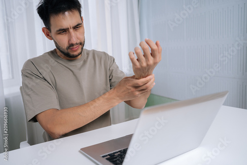 Schilderij op canvas Young man massaging aching wrist at laptop, suffering from carpal tunnel and repetitive strain from prolonged computer use, showing office-related pain and discomfort