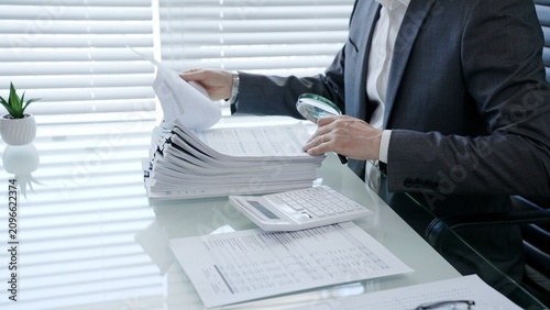Professional businessman using a magnifying glass to meticulously examine financial reports and paper documents on a desk, analyzing data for audit or investigation purposes