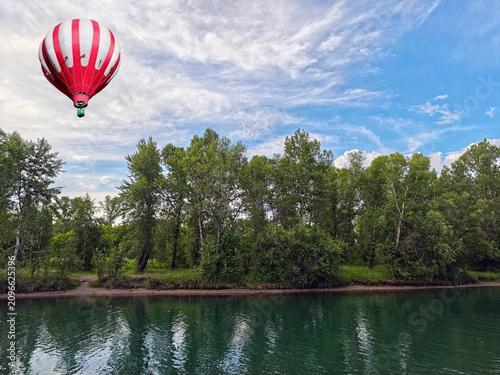 Red and white hot-air balloon floating above the wooded riverbank.