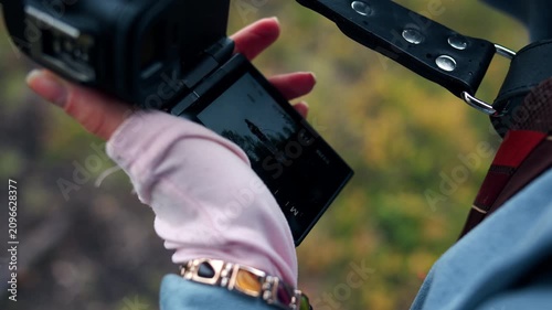 Close-up of a left female hand in a pale pink glove holding a mirrorless camera attached to a leather belt wit flip-out LCD screen tilted toward the viewer. The photographer is taking series of photos