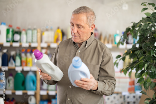 Mature man chooses detergents and liquid laundry gel. Pensioner stands in a store and buys an all-purpose cleaner for surfaces and home