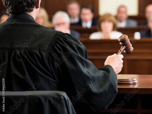 A rear view of a judge wearing a black robe and holding a wooden gavel inside a formal courtroom.