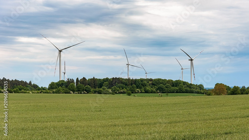 Wind turbines rising above a forest line beyond a green field