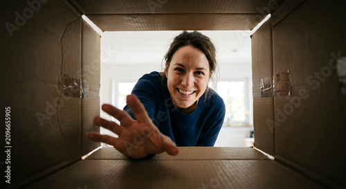 A happy young adult woman reaches into a cardboard box looking at the camera from an inside perspective showing excitement and curiosity. Generative AI.