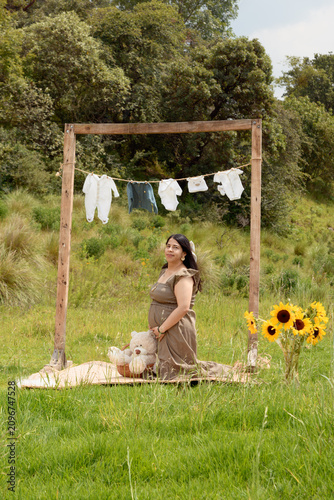 Pregnant Woman Kneeling in Meadow with Baby Clothes Setup