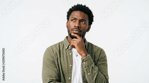 A young black man wearing a green shirt looking up and thinking with his hand on his chin against a plain background