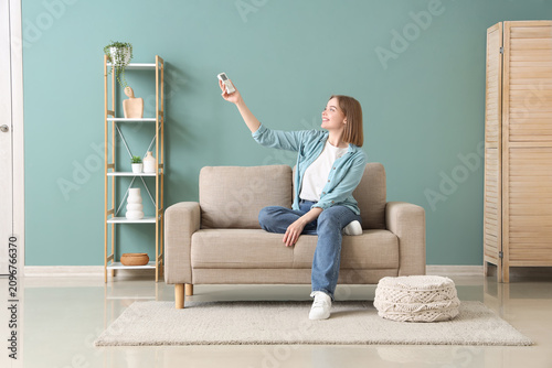 Young woman sitting on a sofa and adjusting the air conditioner with a remote...