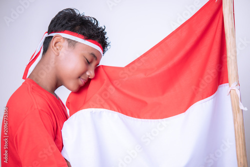 Obraz na płótnie A happy Asian boy celebrating Indonesia's independence day on August 17th stands while kissing the red and white flag, isolated on a white background