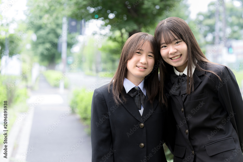 Fototapeta premium 屋外の緑背景で笑顔で寄り添う日本人女子高校生 二人で過ごす高校生活と青春のポートレート カメラ目線の笑顔