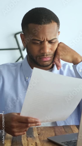 Upset young african american businessman sitting at his desk feeling stressed and disappointed while reading a document with bad news, a notice of dismissal, or a negative business report