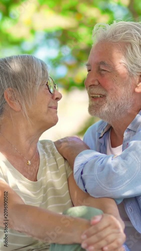 Beautiful senior couple sitting on a park bench, enjoying retirement together. The happy old man with a grey beard laughs and kisses his smiling wife, showing love and affection outdoors