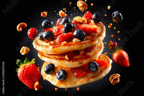 A stack of golden pancakes floating in mid-air topped with strawberries, blueberries, and walnuts, surrounded by scattered nuts and honey droplets against a black background.