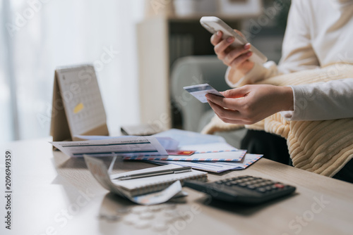 Woman holding credit card and smartphone with unpaid bills on table, representing household debt and extravagant spending lifestyle.