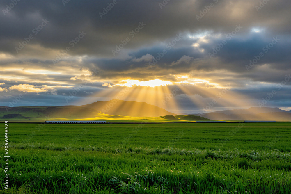 Obraz premium Sunbeams shine through dark clouds over green Icelandic meadows, mountains. Golden light hits grass fields under cloudy sky at dawn. Beautiful rural landscape panorama shows dramatic weather,