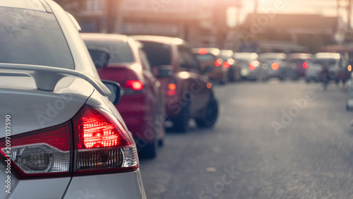 Rear view shot of a silver car waiting in heavy city traffic during the day. The bright red tail light is highlighted as multiple vehicles queue up on the road creating a busy commute scene.