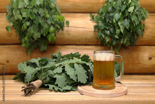 Foto A mug of light beer on a round wooden tray next to a fresh oak broom stands on a plank bench against the background of a log wall with a couple of fresh birch brooms hang on it