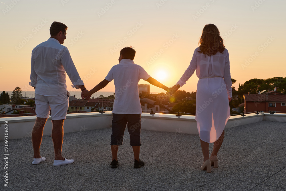 Obraz premium Family Holding Hands on Rooftop Terrace at Sunset With Sea View Over Coastal Town