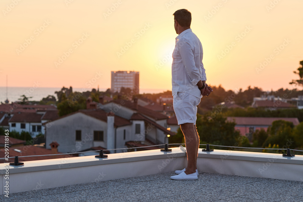 Obraz premium Man Standing on Rooftop Terrace Watching Sunset Over Seaside Town From Home Balcony