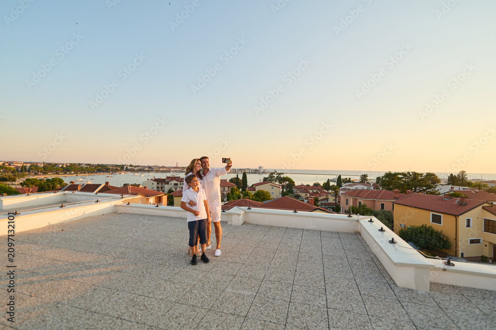 Obraz premium Family Taking Selfie On Rooftop Terrace Overlooking Coastal Town And Sea At Sunset