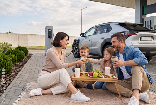 Family Enjoying Picnic Lunch While Electric Car Charges at Modern Roadside EV Station