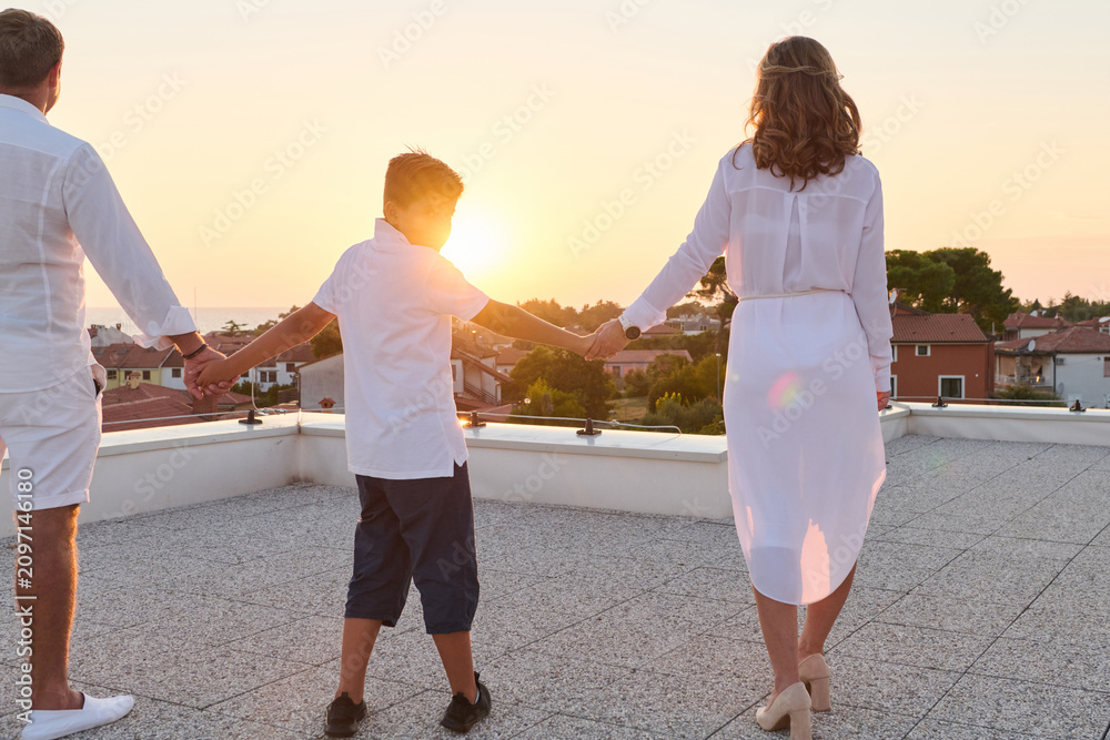 Obraz premium Family On Rooftop Balcony Holding Hands At Sunset With Sea View Over Coastal Town