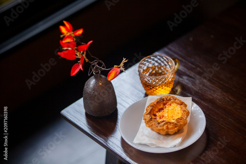 Cozy afternoon tea time with a delicious egg tart served on a white plate, a glass of warm herbal tea, and a decorative small vase with bright red flowers on a dark wooden table.
