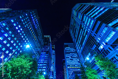 Dynamic low-angle looking up perspective of futuristic corporate skyscrapers lining a dark night sky canyon in Tokyo.