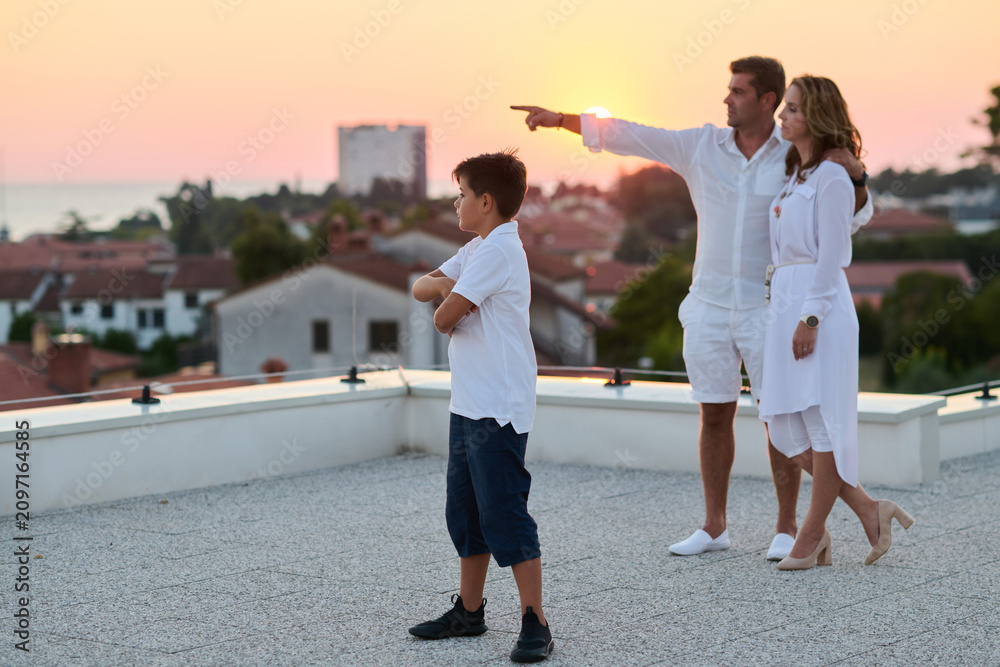 Obraz premium Family On Rooftop Terrace At Sunset With Sea View, Parents Pointing While Son Watches