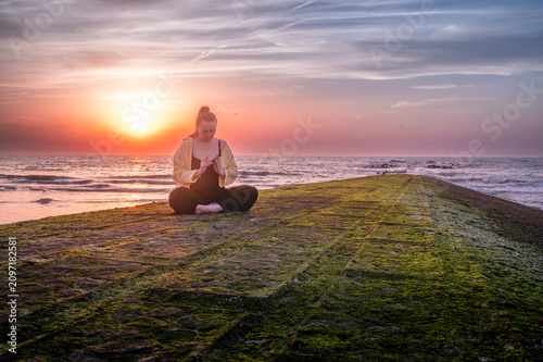 Woman in seated meditation with prayer hands on a moss covered breakwater at sunset a calm and grounding moment of mindfulness and mental clarity beside the sea.