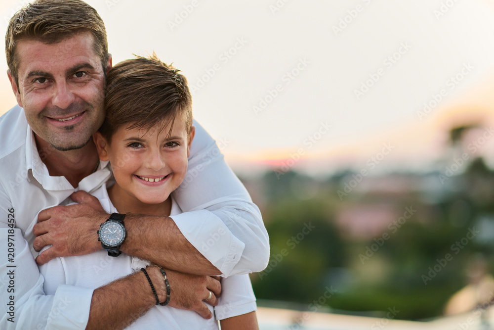 Obraz premium Father Hugging Smiling Son On Balcony At Sunset Overlooking Sea View From Rooftop