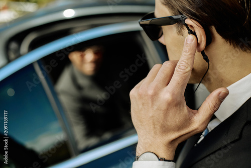Fotografie Close up of young adult Caucasian man adjusting earpiece while guarding client near car, conveying executive protection