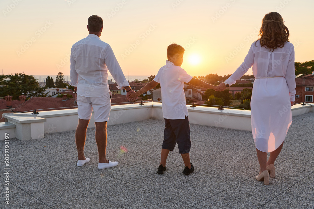 Obraz premium Family Holding Hands on Rooftop Terrace at Sunset With Sea View Over Coastal Town