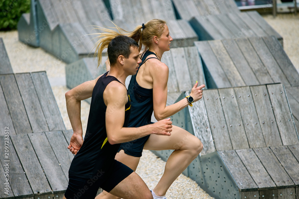 Obraz premium Athletic couple running past modern wooden structures in urban training zone. Concept of architectural sport lifestyle, outdoor partner training, city fitness content, and active wellness campaigns.