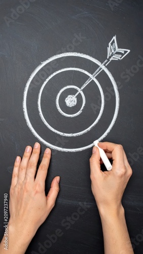 Hands drawing a target with chalk on a dark surface, arrow hitting the bullseye