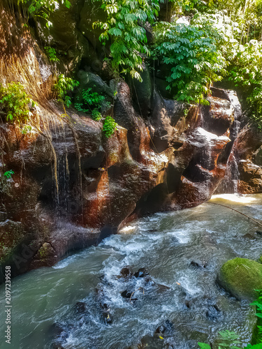 Water streaming down dark reddish rocks into turbulent jungle river with mossy boulders and tropical vegetation