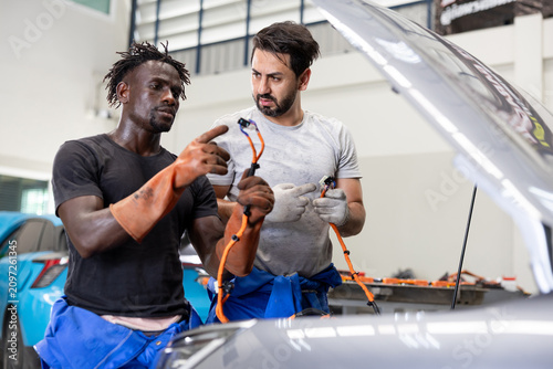 Two auto mechanics examine wiring under an open car hood in a workshop, discussing repairs and diagnostics with greasy hands in a busy garage.