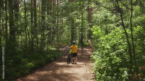 Slow motion rear view of a school age boy walking beside his bicycle and guiding it down a steep forest trail, capturing a calm summer outdoor moment in lush green woodland. High quality 4k footage