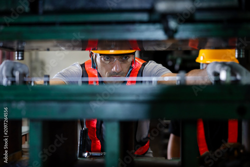 Factory worker in safety gear operating a heavy industrial press, viewed through machinery components, highlighting precision, strength, and workplace safety on the production line.