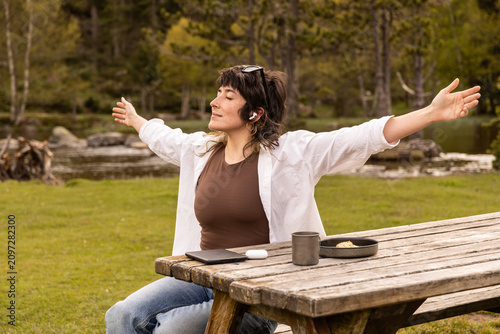 Woman with curly hair sits in a park. Her arm rests on a wooden railing. Sunglasses cover her eyes. Lush green grass and trees surround a small pond. Her calm expression shows peaceful thought